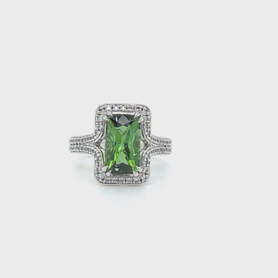 AN EMERALD CUT GREEN TOURMALINE RING SURROUNDED BY A HALO OF SMALL WHITE DIAMONDS WITH SPLIT DIAMOND SET SHOULDERS. THE VIVID GREEN TOURMALINE HAS BEEN SET WITH FOUR CLAWS ANDT THE RING HAS A PLAIN WHITE BACKGROUND